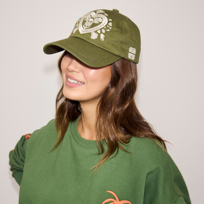 Olive Amara Cap in green cotton with white heart embroidery, worn with a matching green sweatshirt.